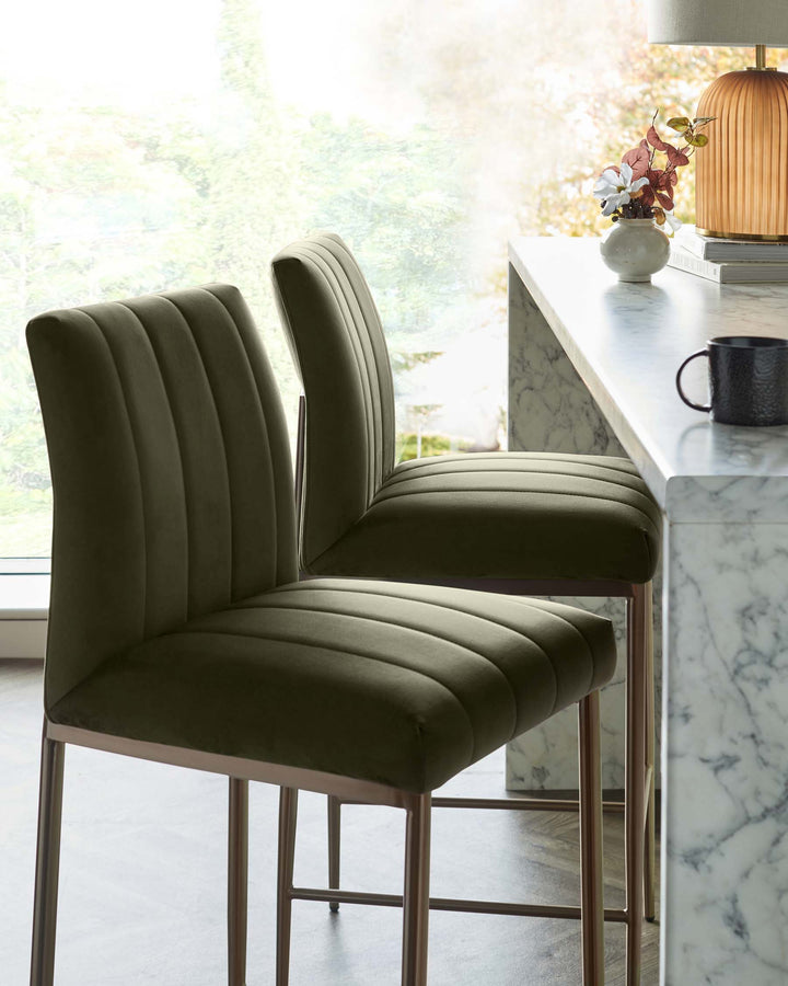 Two stylish bar stools with dark green velvet upholstery and brushed gold legs, set against a bright kitchen backdrop.