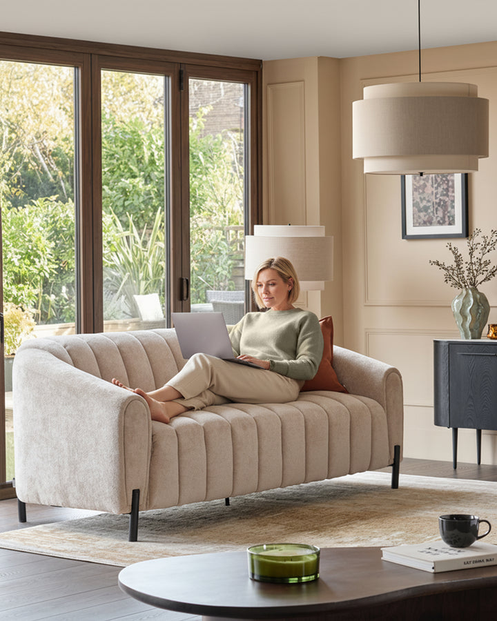 Kendra 3-seater sofa in natural fabric with vertical channel quilting, black metal legs, displayed in a bright living room.