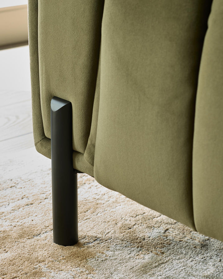Olive green upholstered sofa detail with black metal leg, resting on a neutral patterned rug.