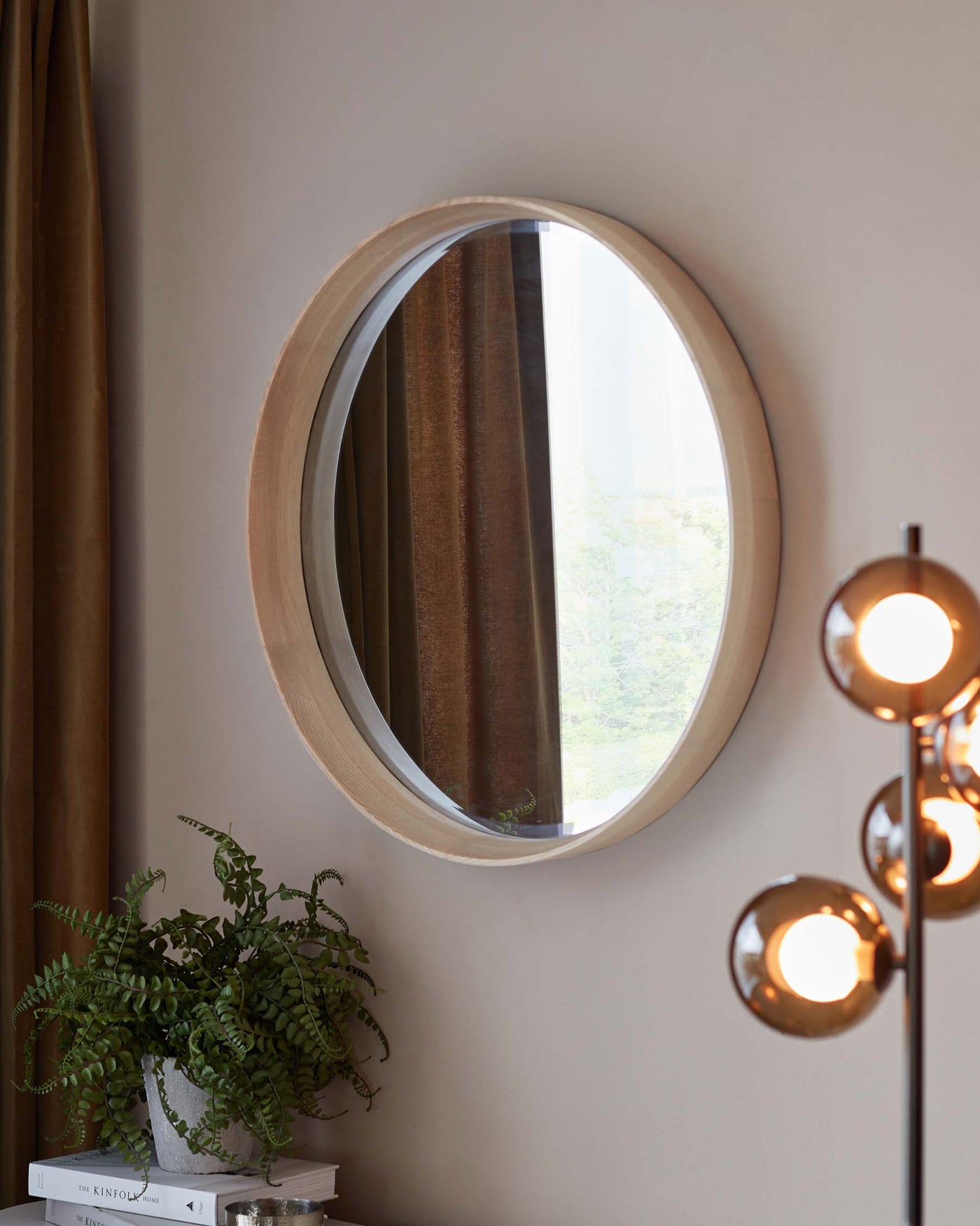 Round wooden mirror with a natural finish mounted on a wall beside a mid-century modern brass floor lamp with multiple globe lights. A potted fern sits on a stack of hardcover books below the mirror, hinting at a cosy reading corner or a stylish entryway vignette.