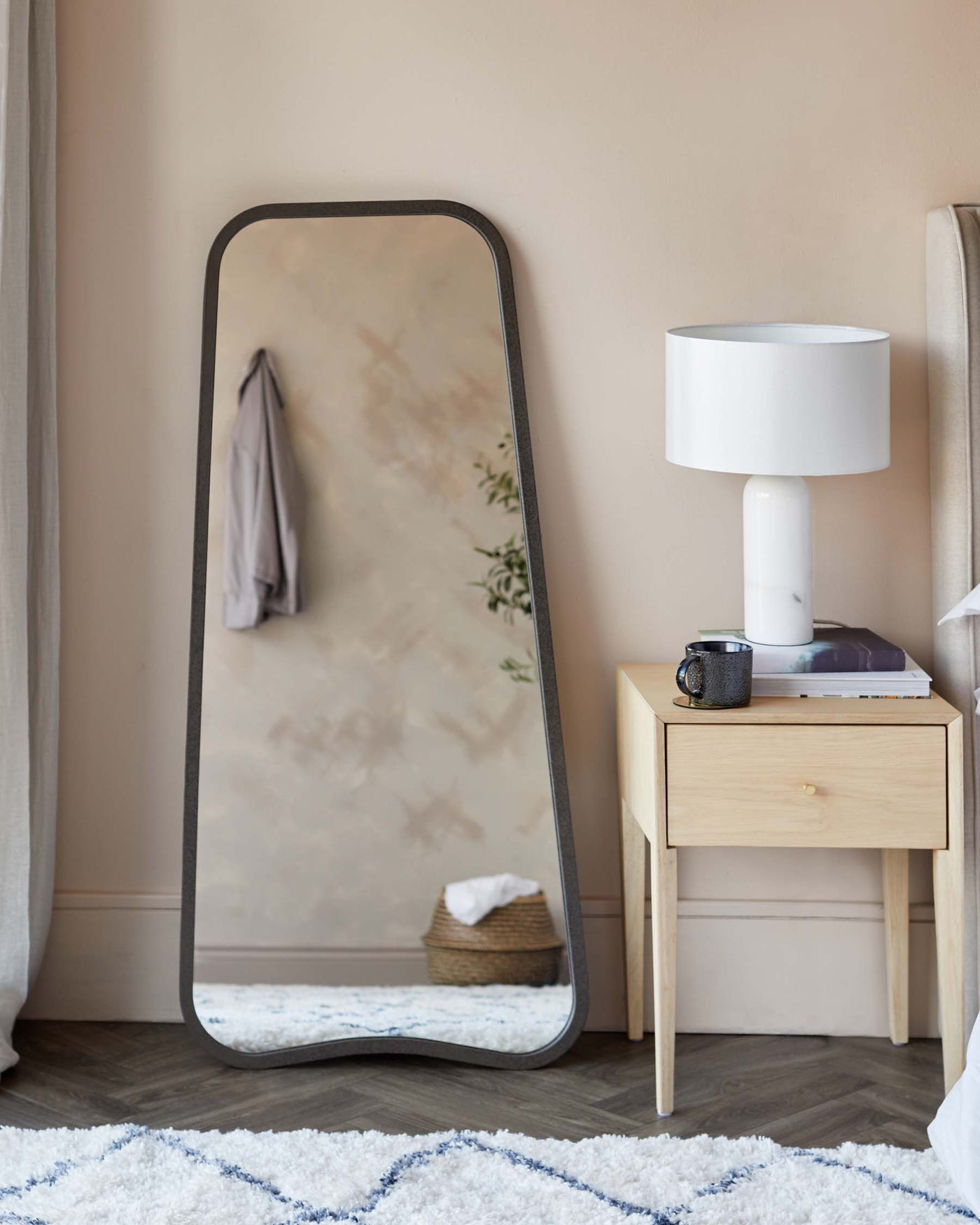 A large freestanding floor mirror with a sleek, curved metal frame, and a minimalist natural wood bedside table with a single drawer and splayed legs, accompanied by a modern white table lamp with a cylindrical marble base.