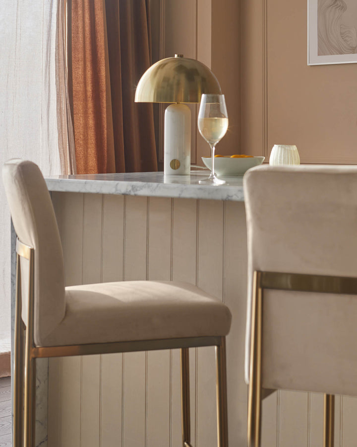 Two stylish bar stools with brass frames are at the kitchen counter, next to a lamp and a glass of wine.