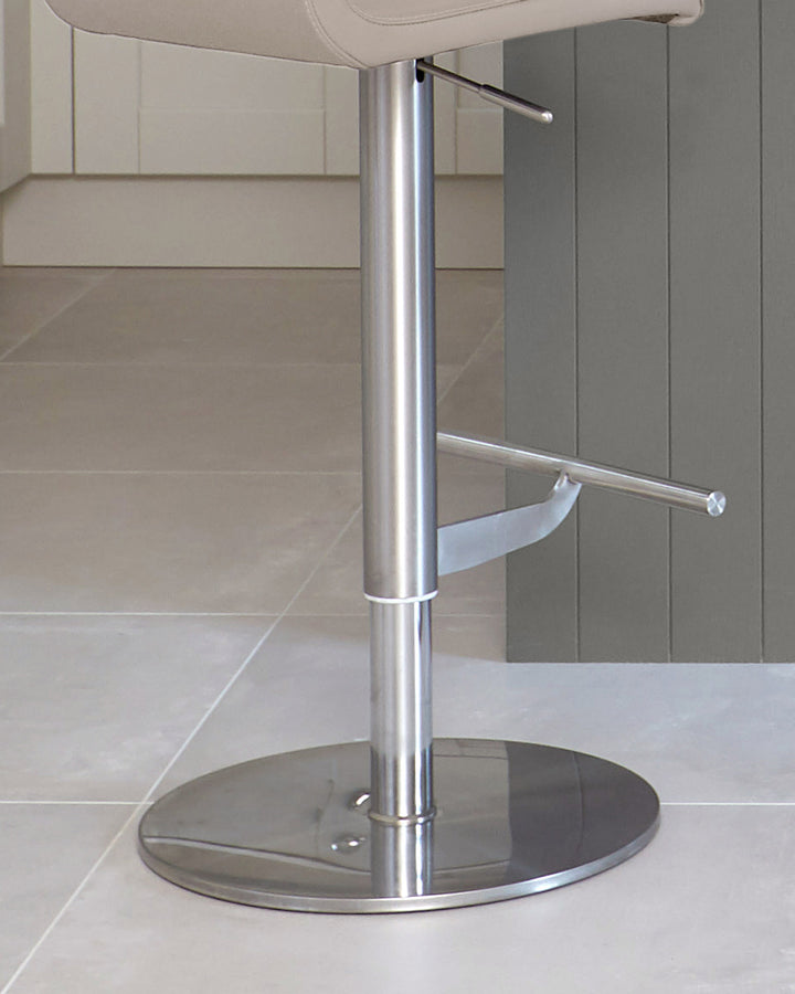 A modern bar stool with a chrome base and a neutral-coloured seat. The stool is in a kitchen setting.