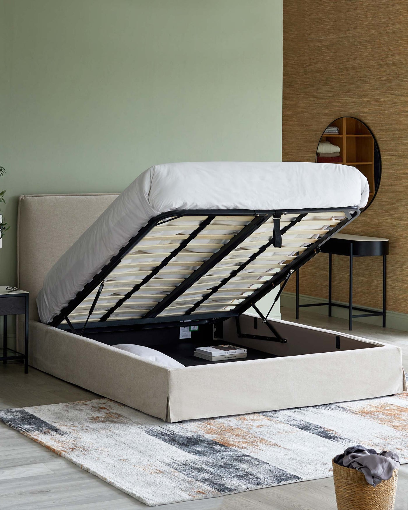 Beige upholstered storage bed with a lifted mattress base revealing a spacious storage compartment, accompanied by a round black side table and a circular wall mirror with a wooden partition, all atop a multi-coloured abstract pattern area rug.