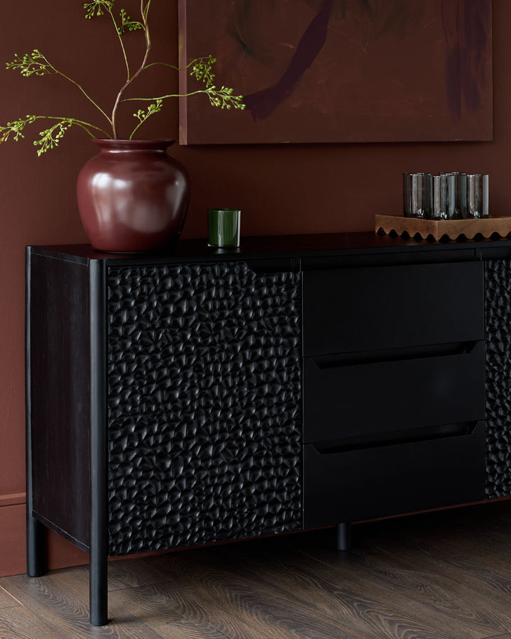 Black wooden sideboard with textured front panels, featuring drawers and doors, displayed against a burgundy wall with decorative vase and greenery.