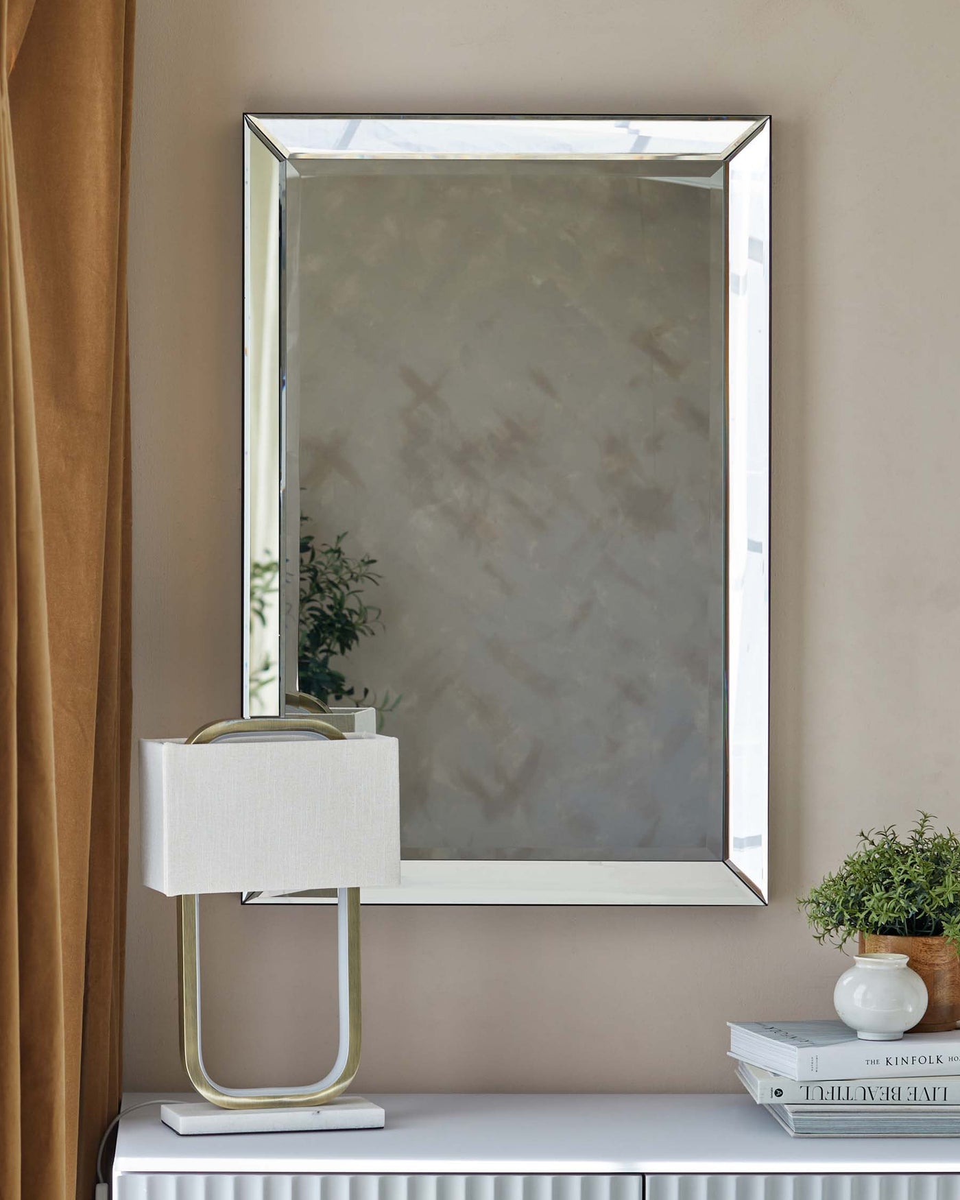 A modern rectangular mirror with a sleek metallic frame mounted on a wall above a white minimalist console table with a stack of books and a potted plant. Beside the table, a contemporary lamp with a gold-finish metal base and a rectangular fabric shade provides an accent to the space.