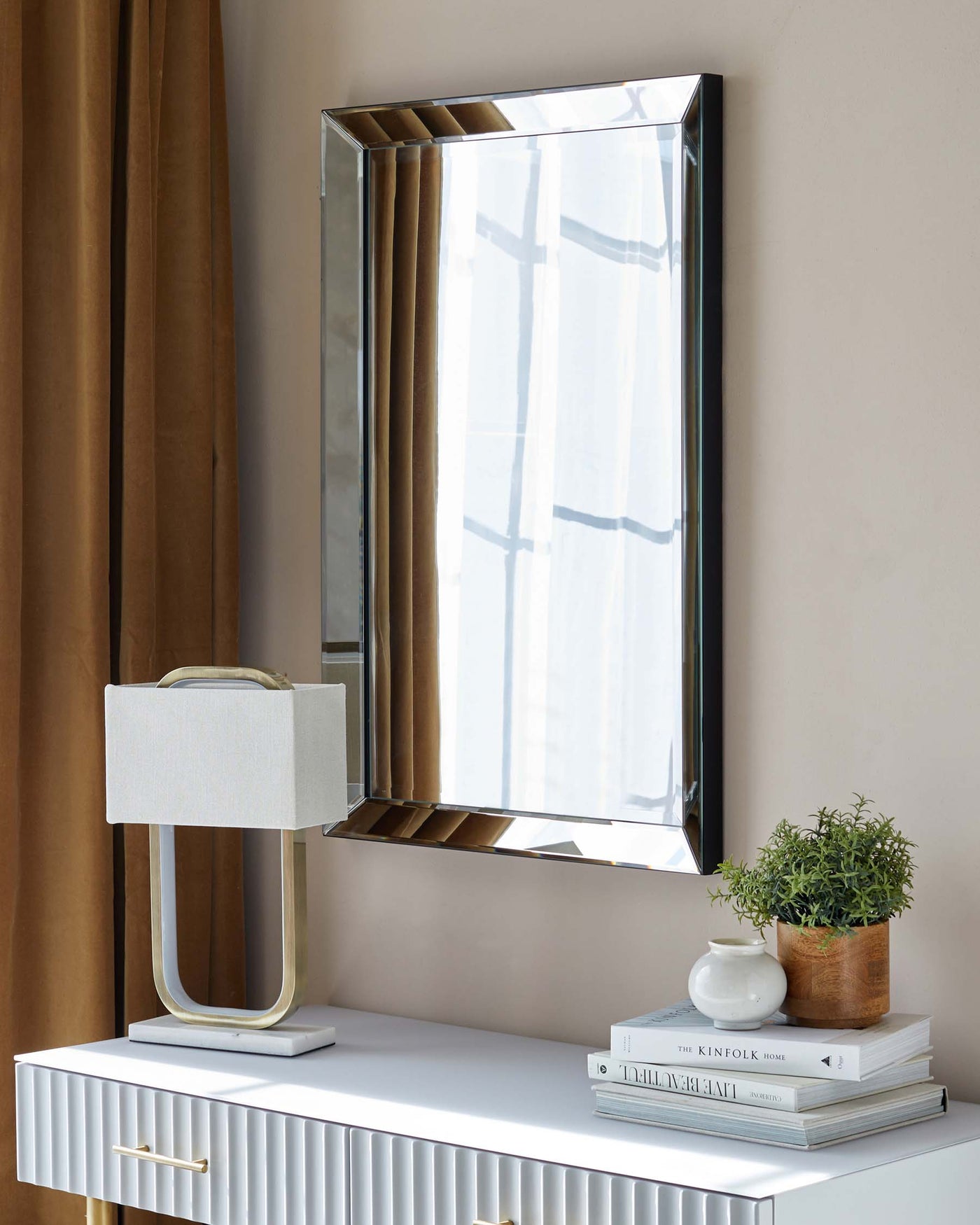 White modern console table with ribbed drawers and brass handles, topped with a contemporary lamp and stacked books beside a potted plant. A rectangular mirror with a layered frame hangs above.