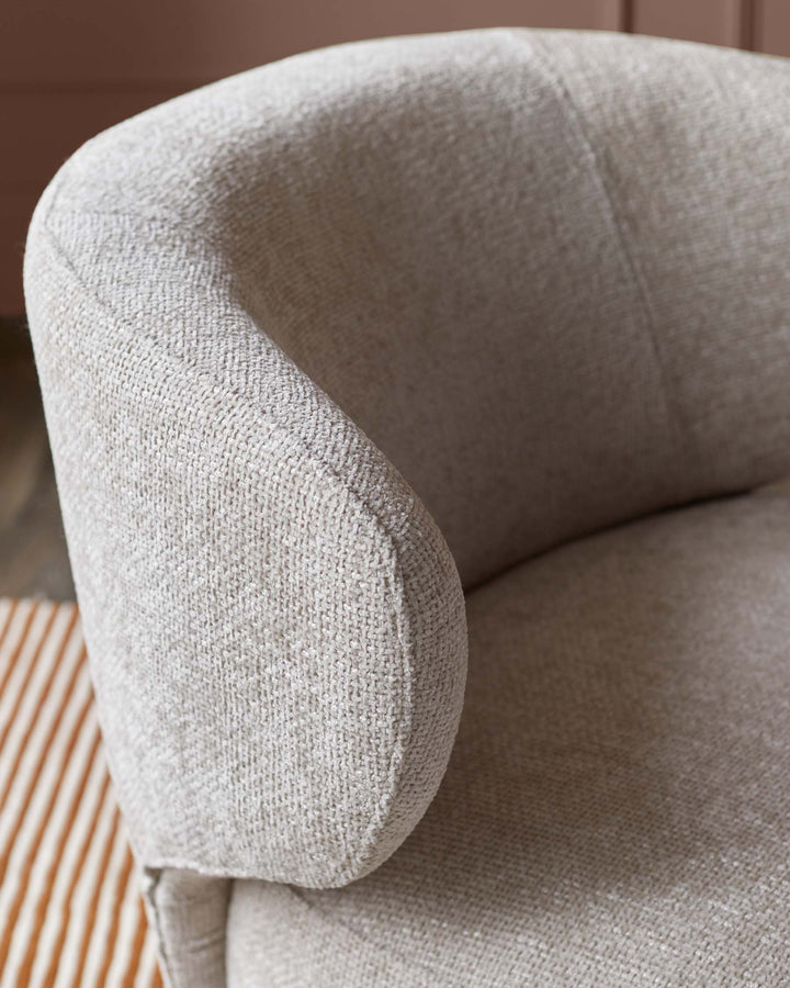 Close-up of a curved, light grey fabric armchair. Soft, textured material with visible stitching along the armrest.