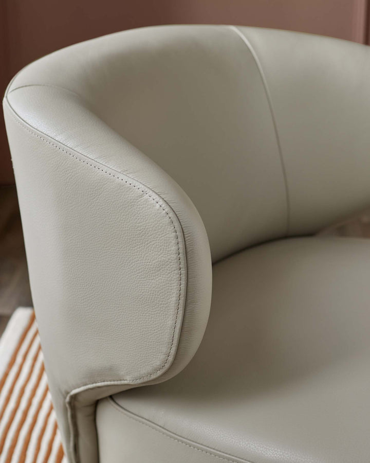 Close up of a grey leather chair with a curved armrest and visible stitching detail, against a pale pink wall and striped rug.