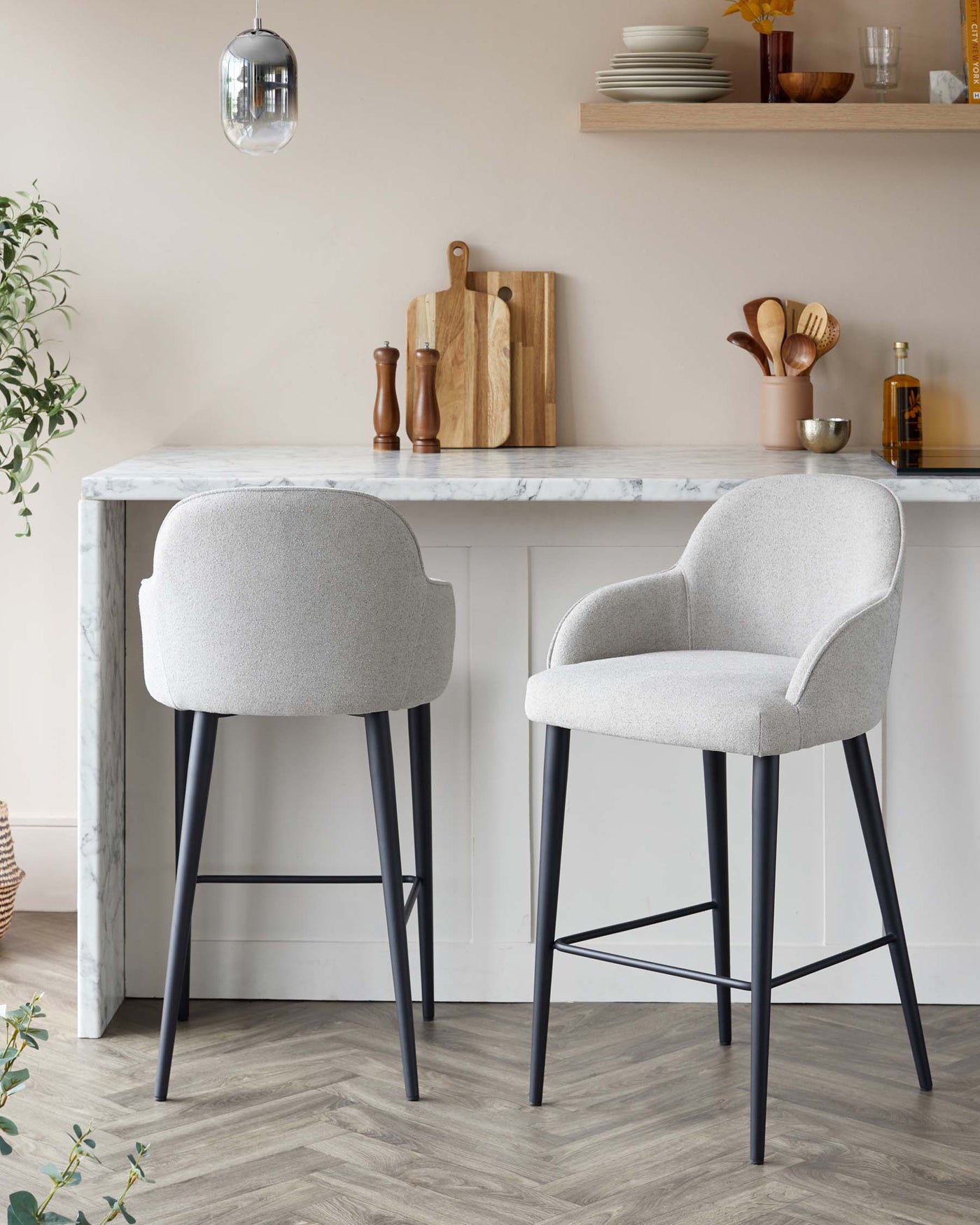 Two modern upholstered bar stools with sleek grey fabric and black metal legs are positioned at a white marble countertop. The stools feature curved backrests and integrated footrests for comfort and style.