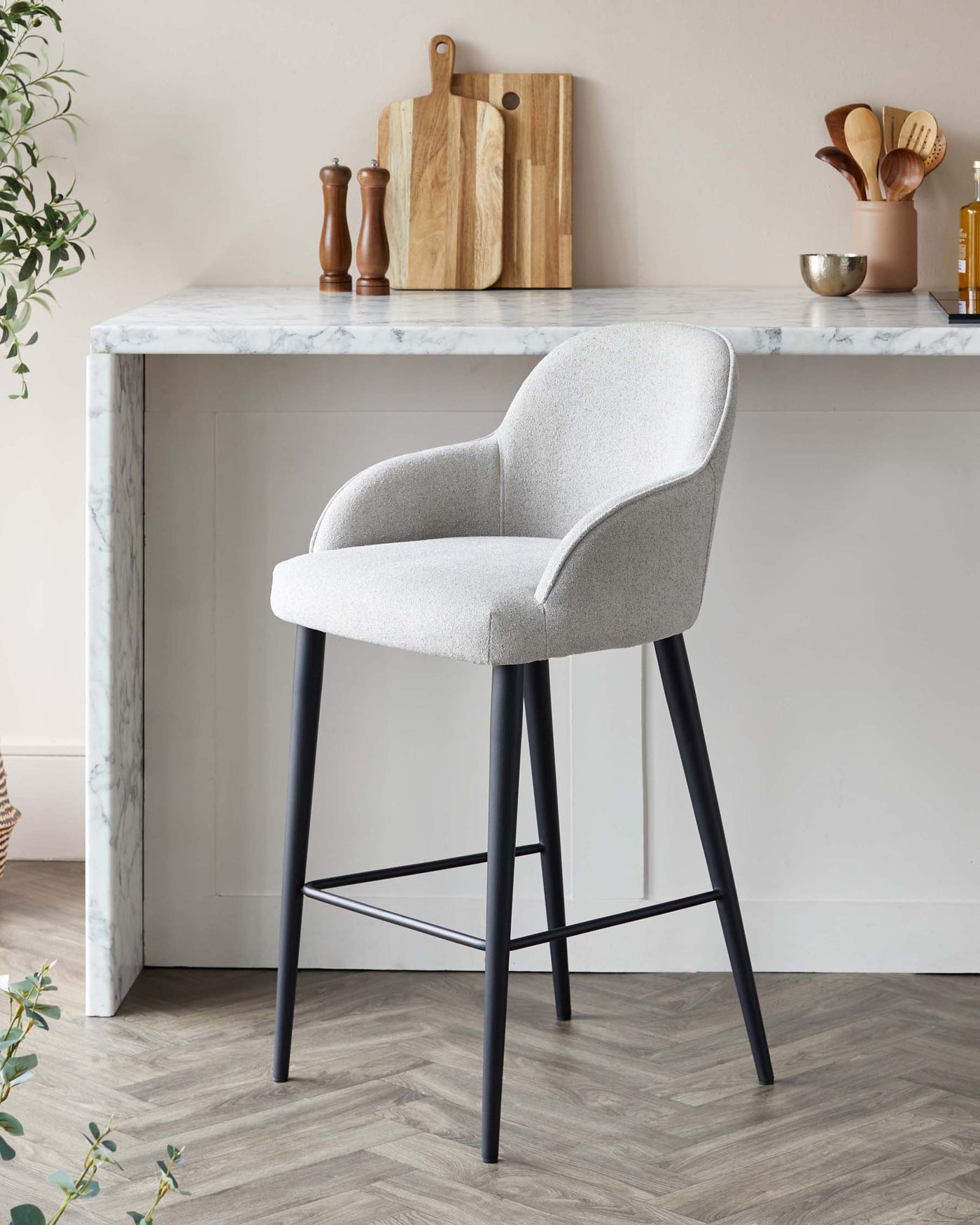 Elegant, modern bar stool with a curved backrest and plush light grey upholstery, featuring slender black metal legs with a footrest, complementing the sleek marble countertop of a minimalist kitchen island.
