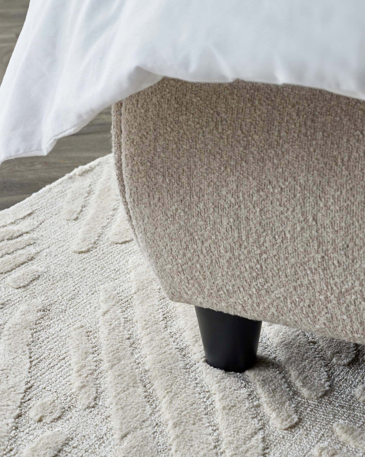 Close-up of a contemporary bed footboard with a textured beige upholstery and a tapered black leg, partially draped with a crisp white sheet, set on a plush cream-colored rug with a subtle geometric pattern.