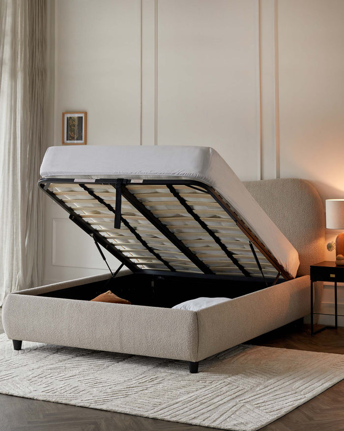 Contemporary upholstered storage bed with a tufted headboard, featuring an easy-lift hydraulic system that exposes ample under-bed storage. Shown in a neutral fabric with a textured finish, complemented by dark tapered wooden legs. Set on a cosy textured area rug in a softly lit bedroom ambiance.