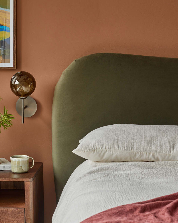Bedroom with olive-green headboard, timber bedside table, globe light and white bedlinen with a pink throw.