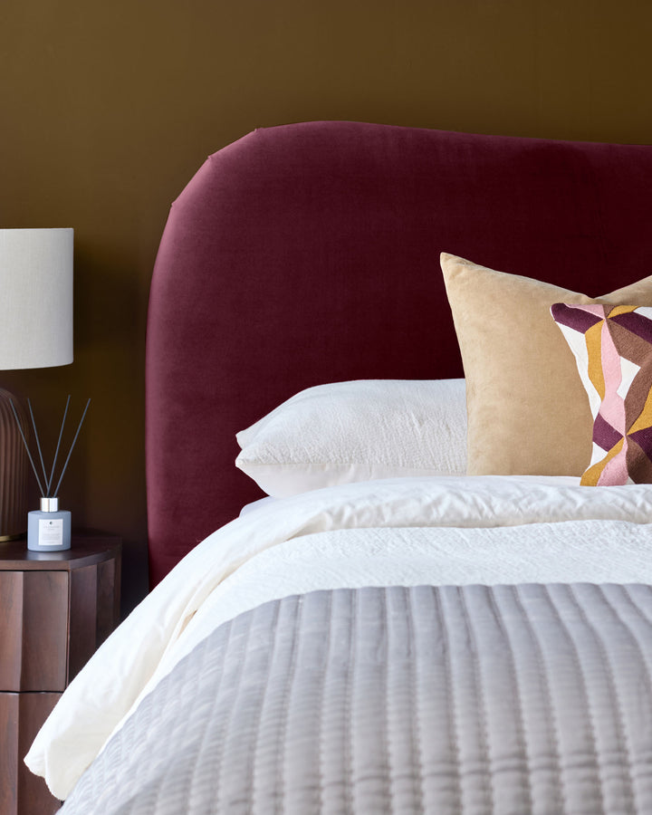 Lani King Ottoman Bed in plum velvet with curved headboard, white bedding, and decorative patterned pillow.