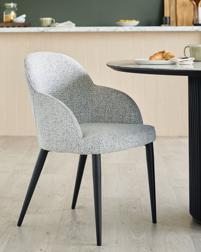 Upholstered chair with black legs and round table with food and drink on top.