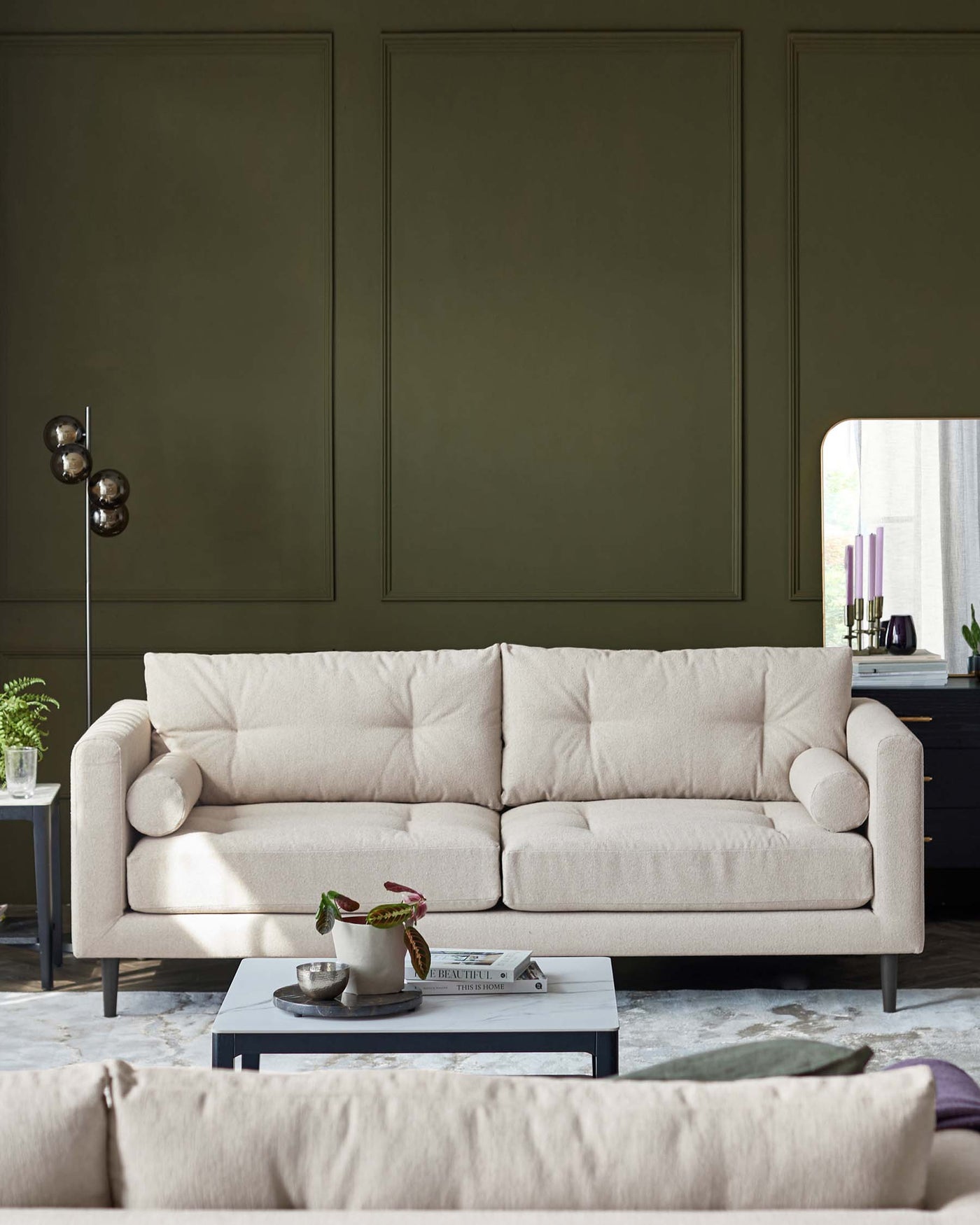 A contemporary beige three-seater sofa with plush cushions and a cylindrical bolster pillow on one side, featuring minimalist wooden legs and subtly rolled armrests, is positioned in front of a muted green wall with decorative panelling. The sofa is complemented by a low-profile grey rectangular coffee table with a book and a small tray containing a decorative plant and candle, all atop a patterned area rug. A modern floor lamp with a metallic finish and spherical shades stands to the left.