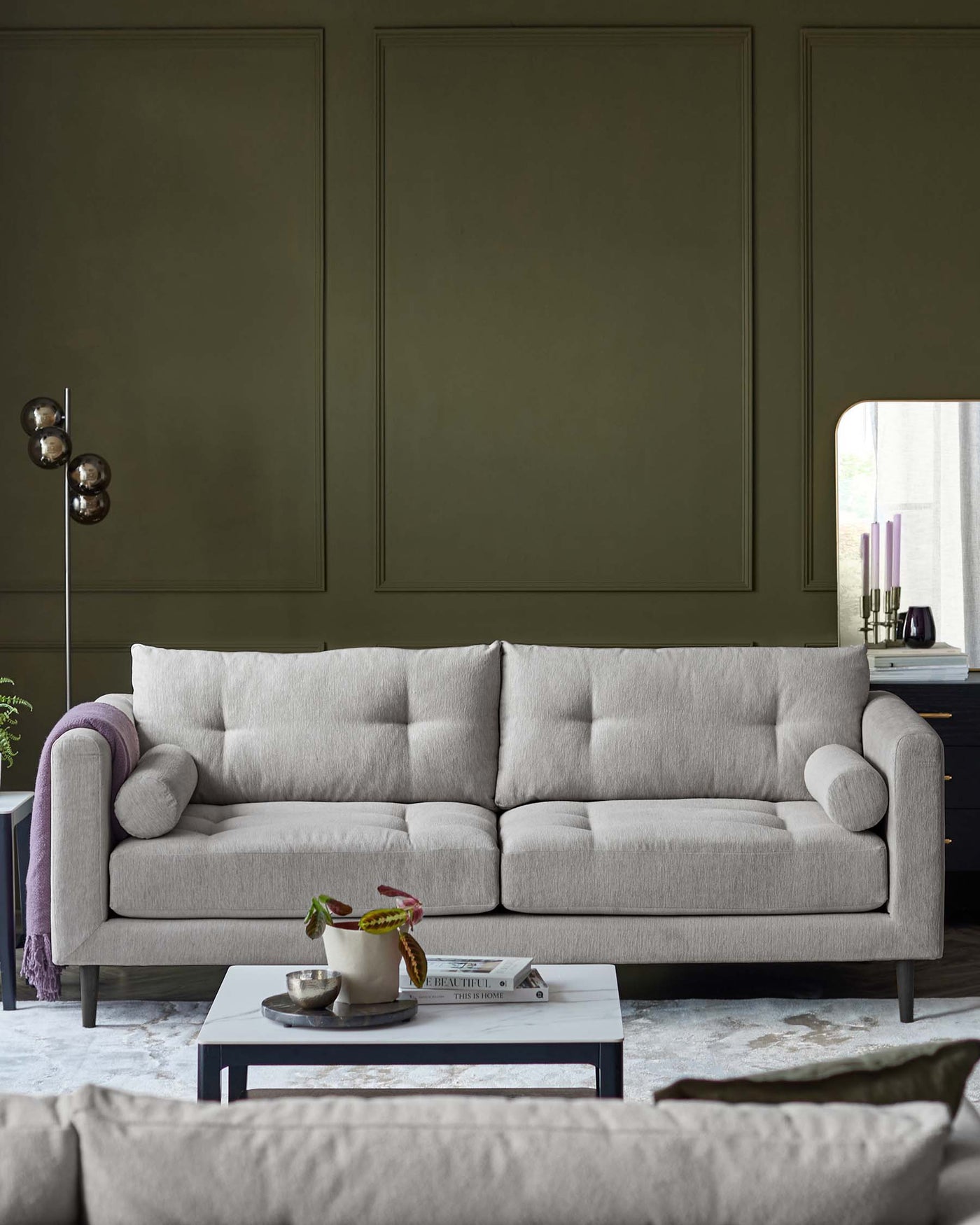 A contemporary three-seater sofa upholstered in a light grey fabric with plush seating cushions, rounded armrests, and cylindrical lumbar pillow. In front of the sofa is a rectangular, dark grey coffee table upon which rests a decorative bowl with plant, a book, and an ornamental object.