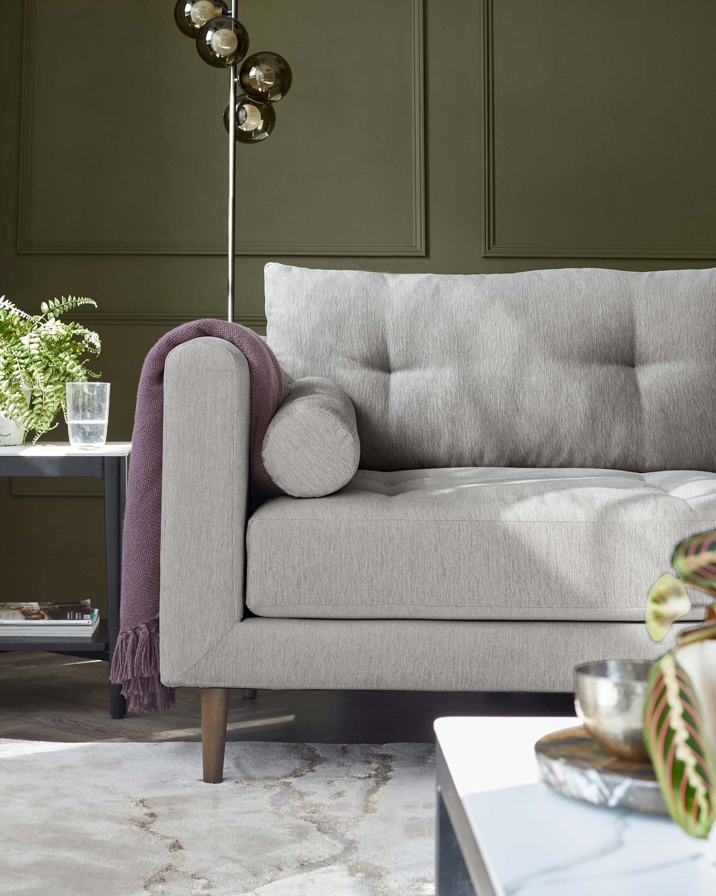 Modern grey upholstered sofa with wooden legs, two cushions, and a round bolster pillow, paired with a purple throw blanket over the armrest. A minimalist glass side table with metallic bowls and greenery is partially visible in the foreground, all set against a muted green wall and atop a textured grey area rug.