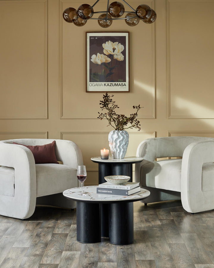 A cosy lounge featuring two modern armchairs, a round side table, stylish lighting and wall art in neutral tones.