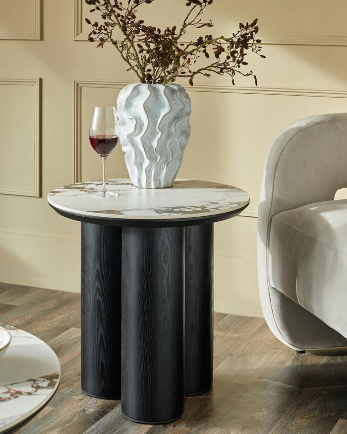 A marble-topped side table with a dark wood base, adorned with a vase and a glass of red wine, beside a modern armchair.