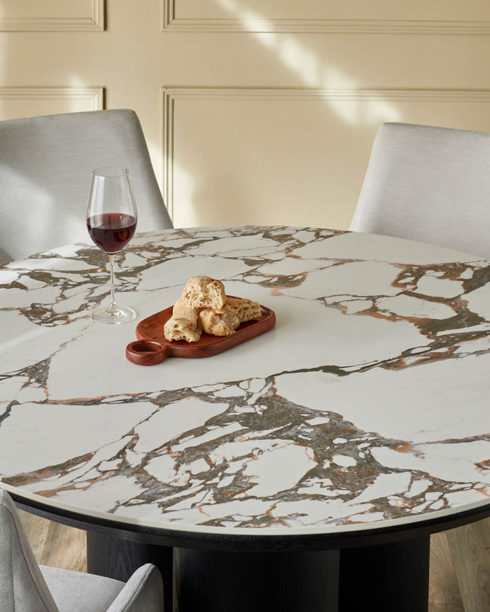 A round marble table with chairs, a glass of wine, and bread on a wooden board are on display.