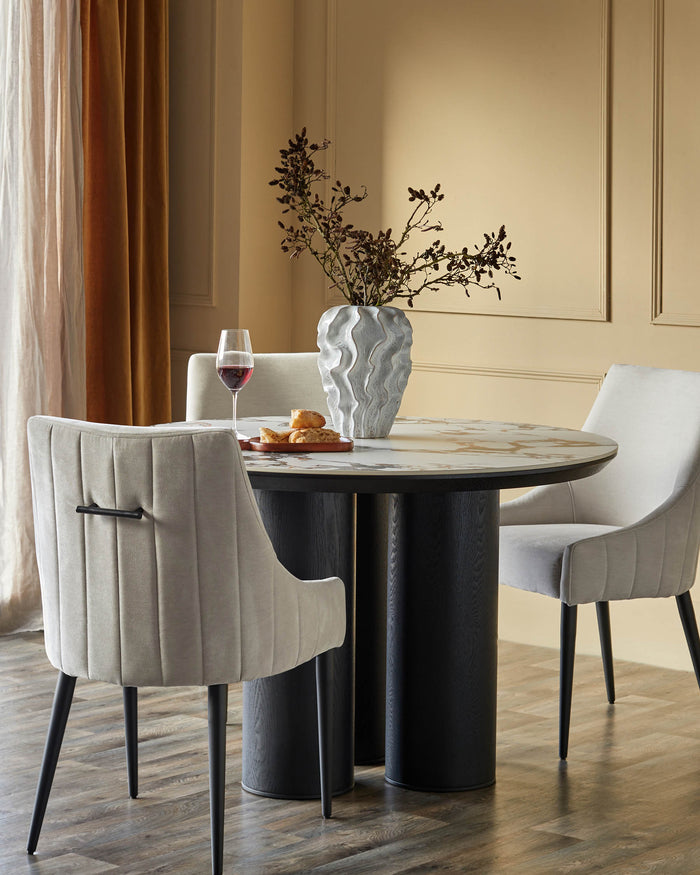 A round dining table with light grey velvet chairs, featuring a decorative vase and a glass of red wine.