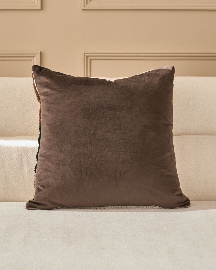 Brown velvet square cushion with fringe trim displayed on a cream-colored sofa against a beige paneled wall.