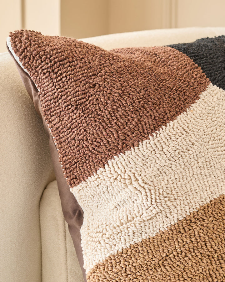 Lennon cushion in natural tones featuring horizontal stripes of brown, cream, and tan textured fabric, displayed on a beige sofa.
