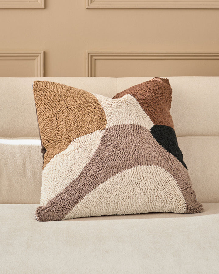 Lennon medium cushion in natural tones featuring geometric color-blocked design in cream, tan, brown and black on a beige sofa.