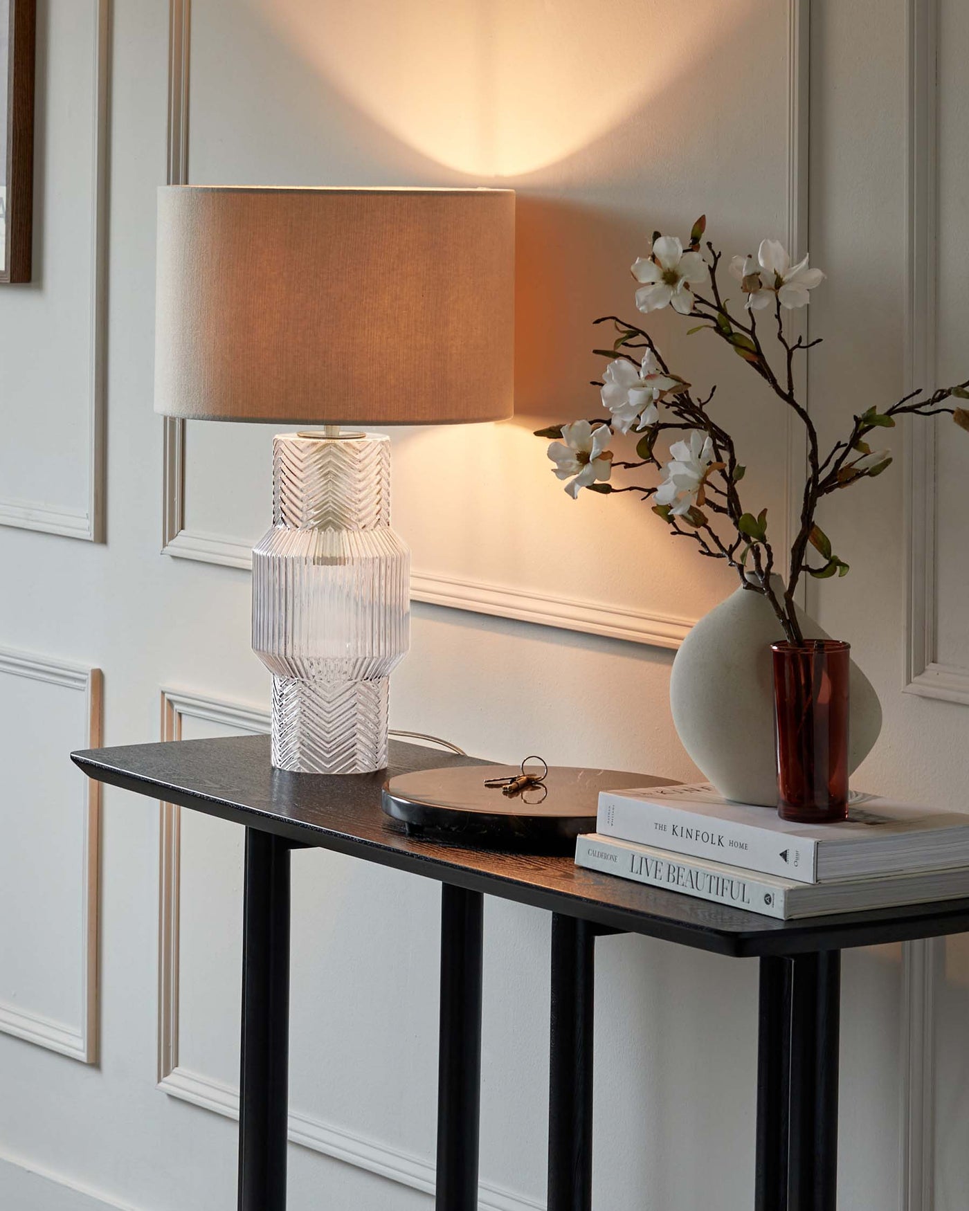 Elegant console table with a dark finish and straight legs, topped with a textured table lamp, a vase with white blossoms, and a stack of decorative books.