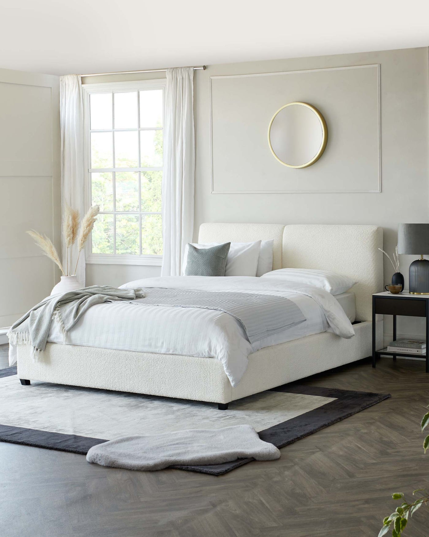 Modern minimalist bedroom featuring a large king-sized bed with a textured white headboard and a white and grey bedding set. A simple, round gold-framed mirror hangs above the bed. To the right, there is a sleek two-drawer nightstand with a grey lamp and books, showcasing a balance of aesthetics and functionality. The room is complemented by a layering of two area rugs beneath the bed, adding texture and warmth to the space.