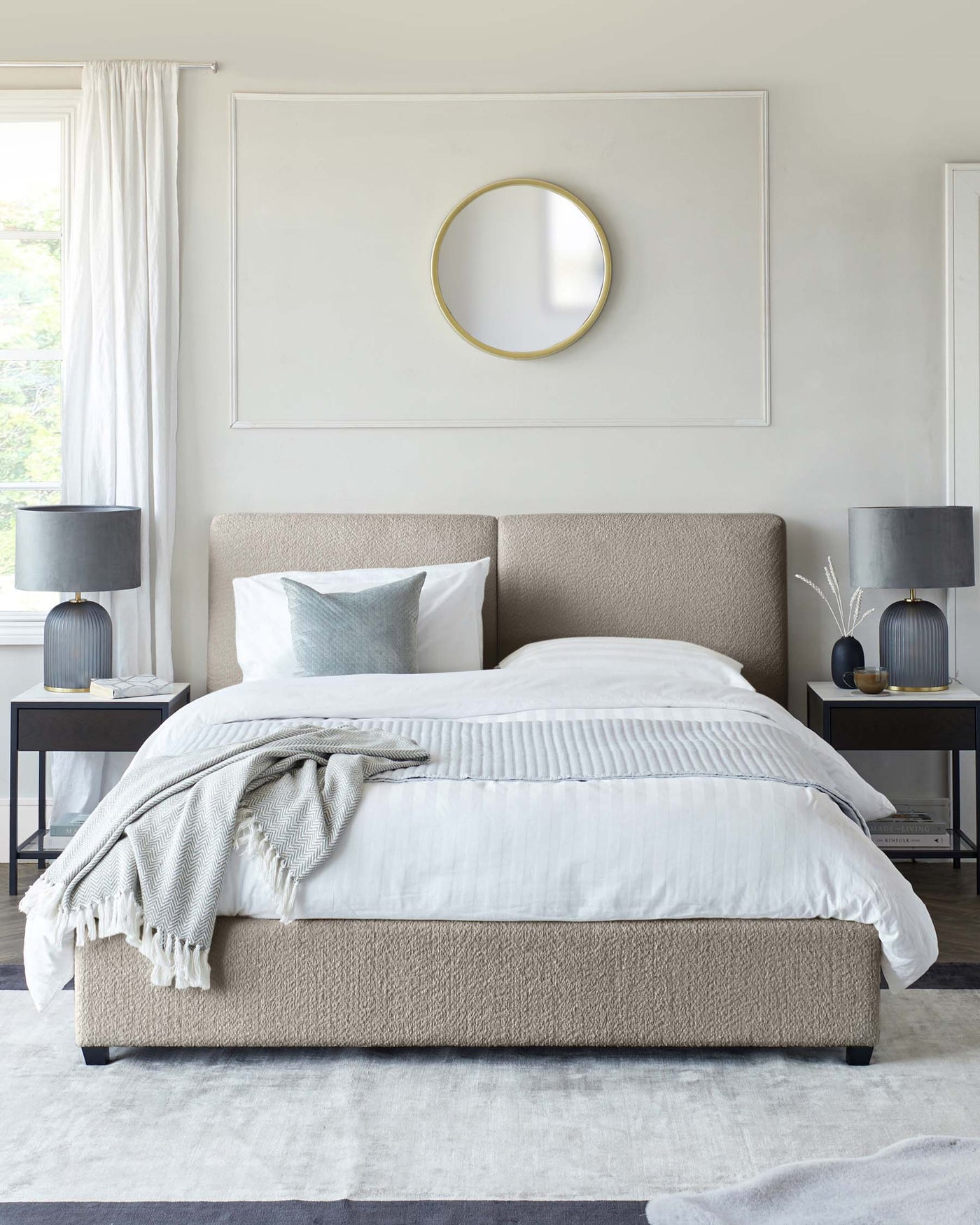 Elegant modern bedroom featuring a lightly textured tan upholstered platform bed with a simple headboard, crisp white bedding, and a soft grey throw blanket. Flanking the bed are two matching black bedside tables with round golden knob detailing, each supporting a texturally ribbed black table lamp. A minimalist round gold-trimmed mirror punctuates the wall above the bed, and the room is anchored by a large light grey area rug, creating a serene and sophisticated ambiance.