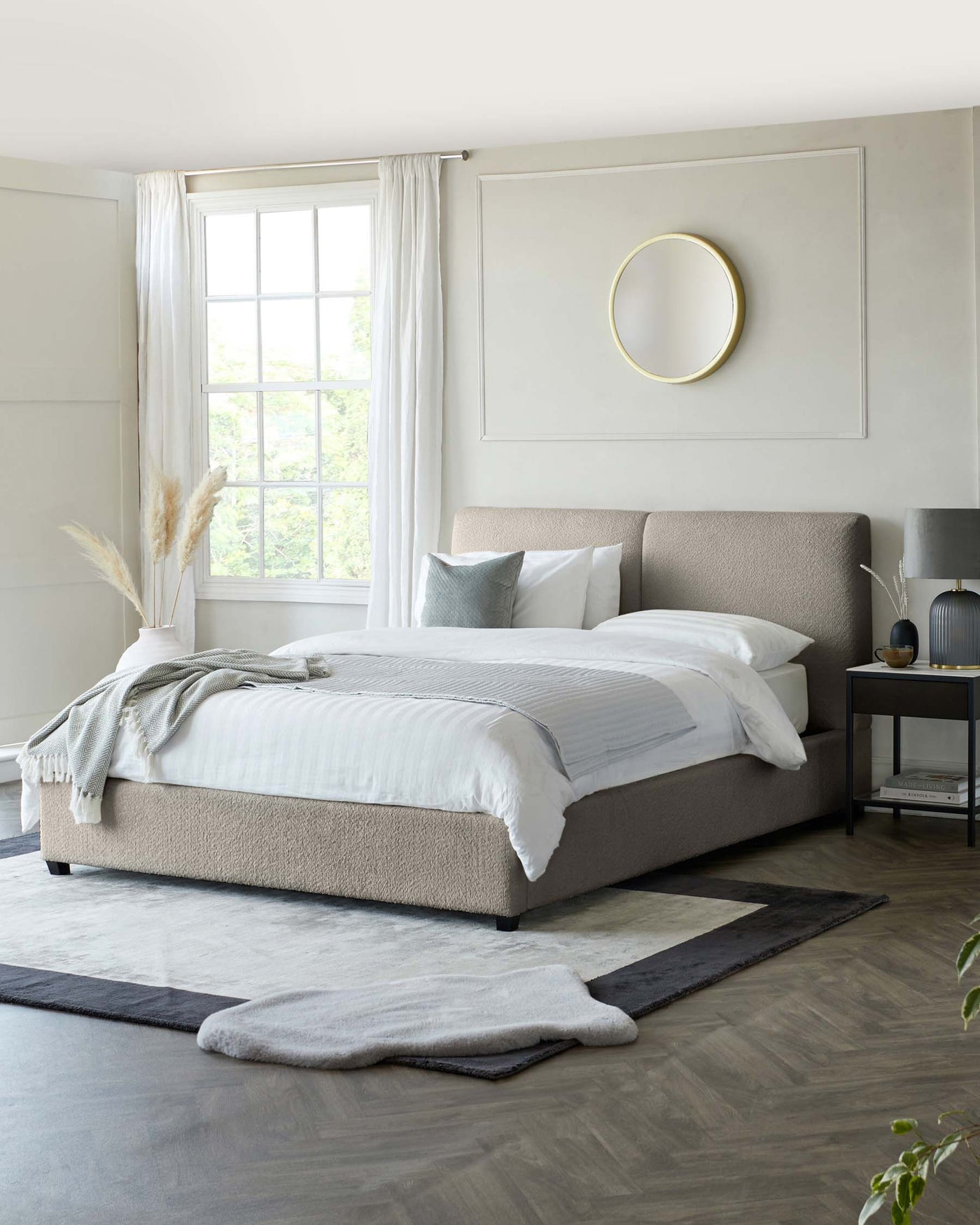 Modern minimalist bedroom featuring a large upholstered platform bed in a neutral beige tone with a textured headboard, crisp white bedding, and assorted grey accent pillows. A contemporary nightstand with a dark finish sits beside the bed, topped with a sleek table lamp and decorative items. A layered area rug combination under the bed adds depth to the room's design.