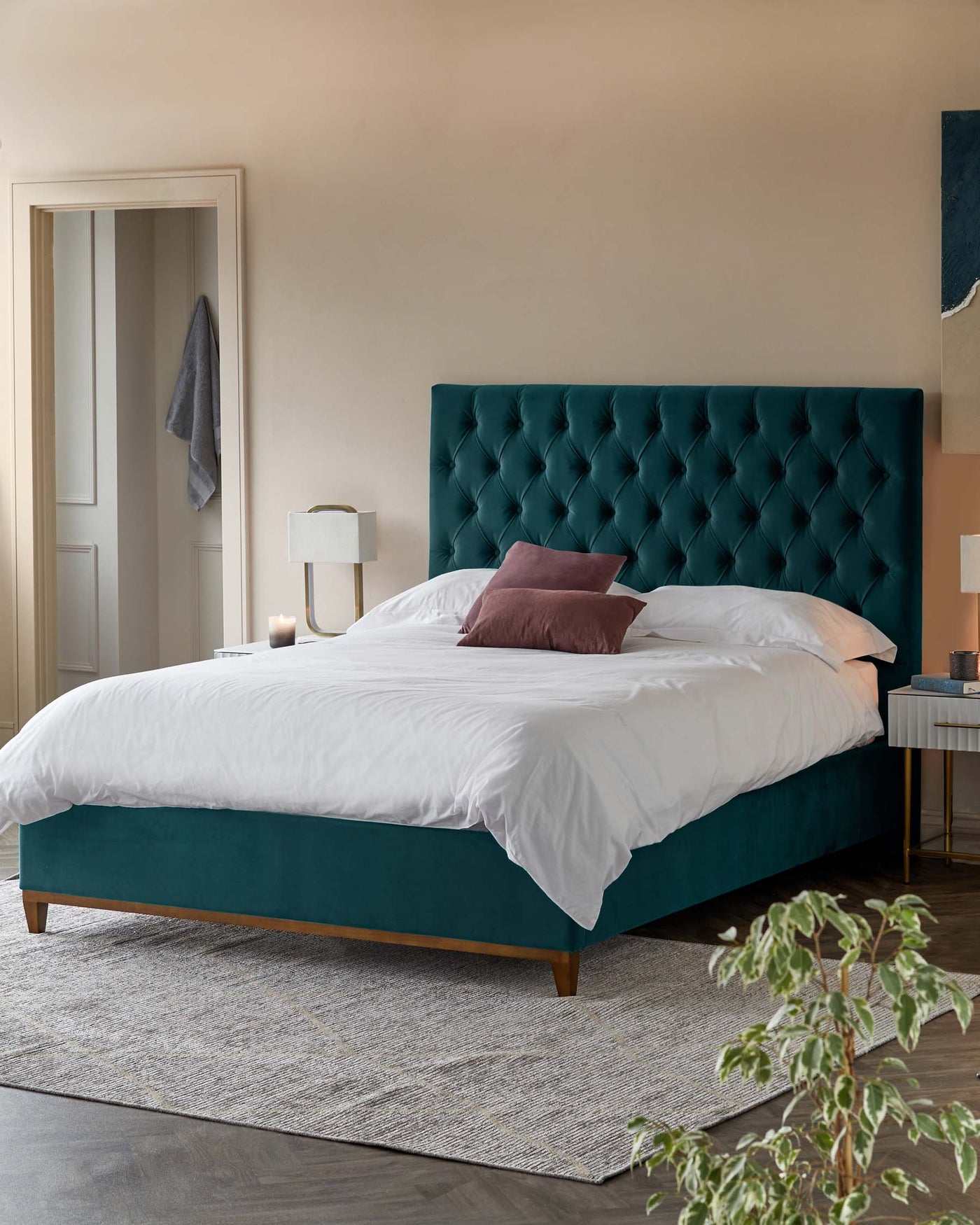 A luxurious teal upholstered king-size bed with a tall, tufted headboard. The bed is dressed in crisp white bedding with a single rose-coloured accent pillow. On the right side of the bed stands a modern bedside table with a white marble top, accented with a gold base. Atop the table sits a white cylindrical table lamp alongside a small decorative item. Below the bed lies a large textured area rug in shades of grey and beige.