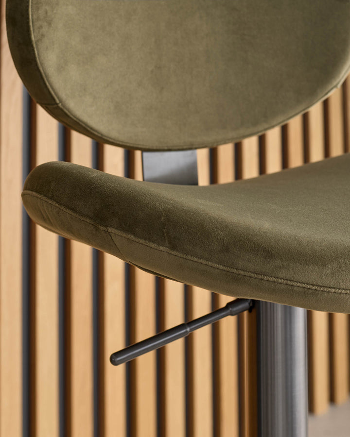 Lilah olive green velvet bar stool with black metal armrest and gas lift base, shown against slatted wood background.