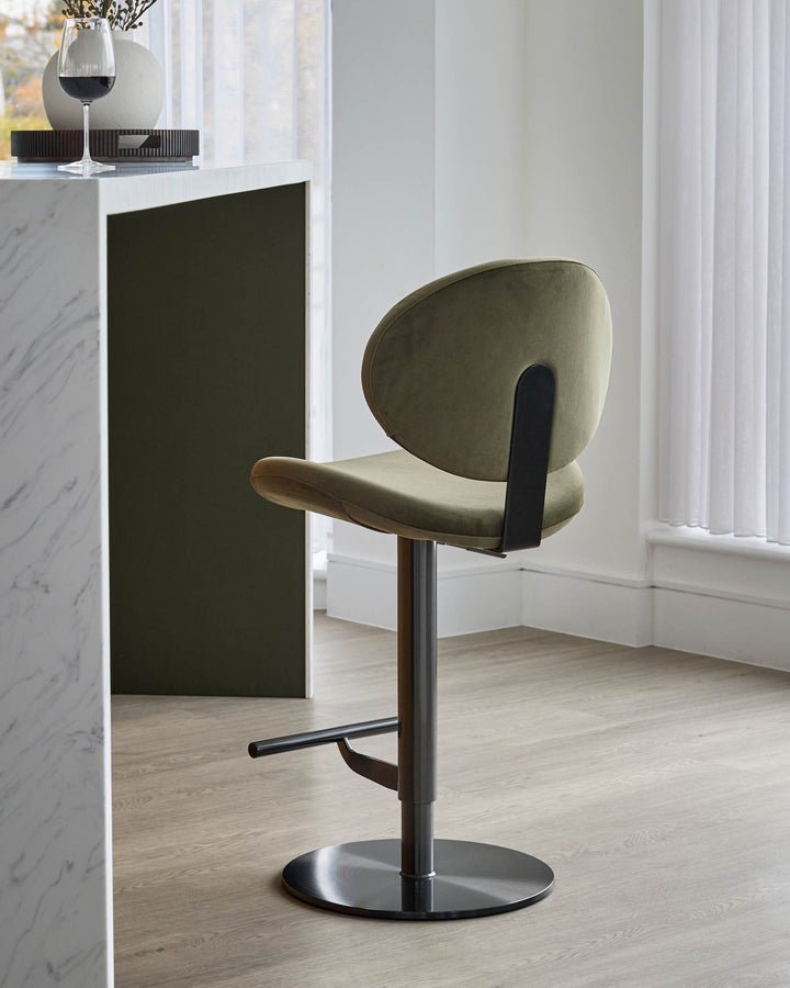 Lilah gas-lift bar stool in olive-green velvet with black metal base and footrest, shown in modern interior setting.