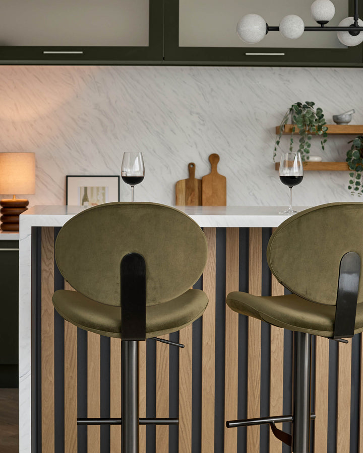 Lilah gas lift bar stool in olive green velvet with black metal frame, shown at kitchen island with marble backsplash.