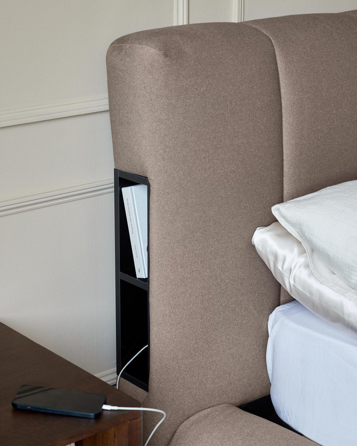 Upholstered bed with built in bedside storage and a dark wood nightstand with a smartphone on top.