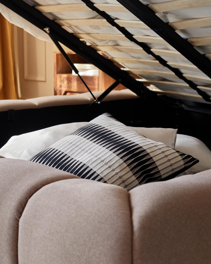 An open ottoman bed with a black frame and a beige fabric headboard and base with a striped pillow on top.