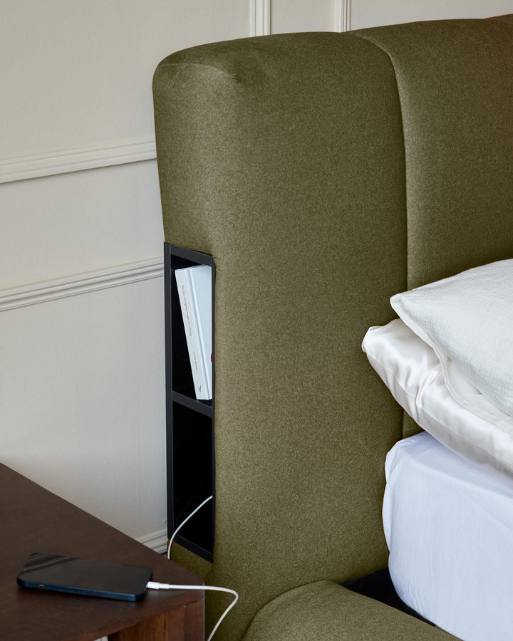 Olive-green upholstered headboard with integrated bedside storage compartment and black frame detail, shown with white bedding.