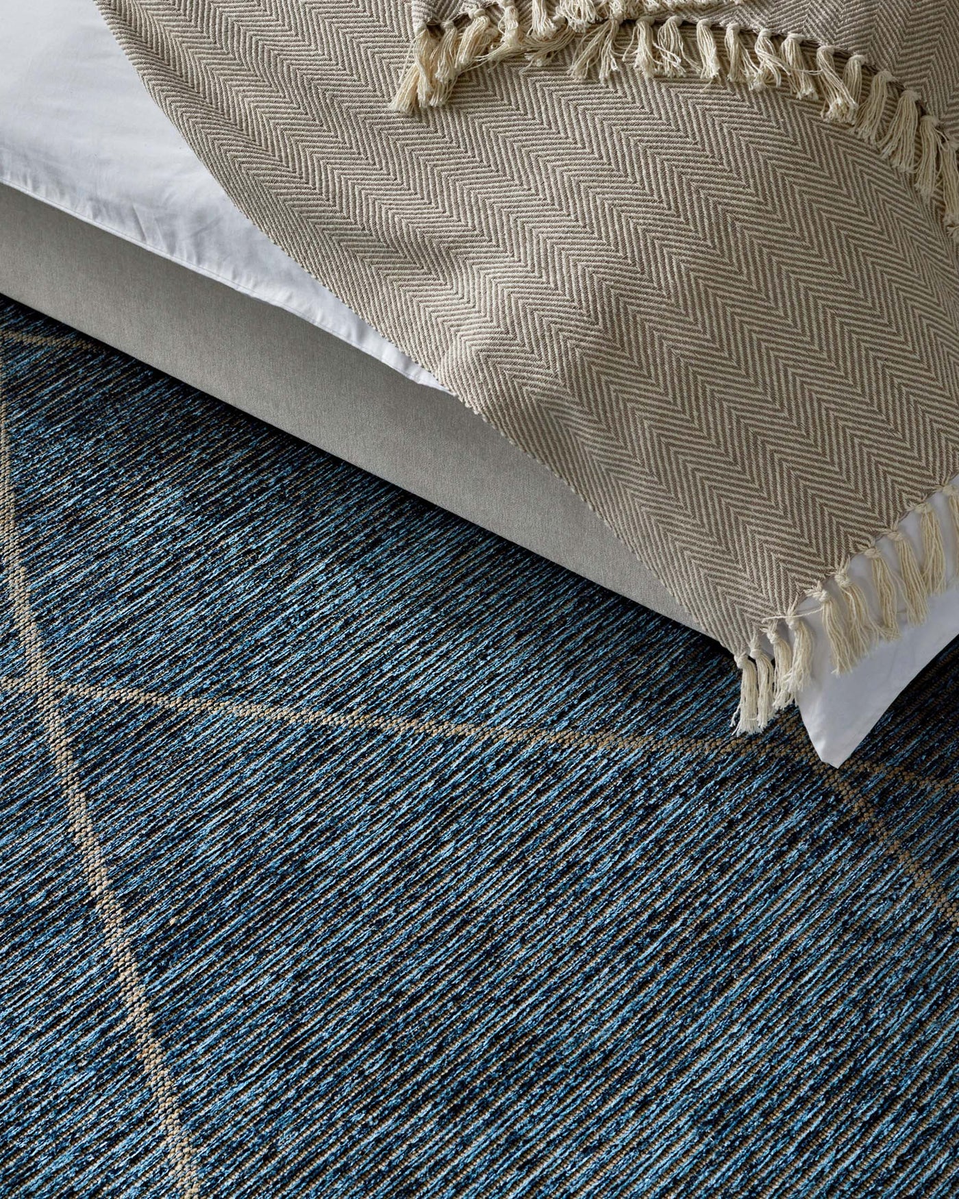Corner of a minimalist upholstered platform bed with a herringbone-patterned throw blanket atop, set against a textured blue and black area rug.