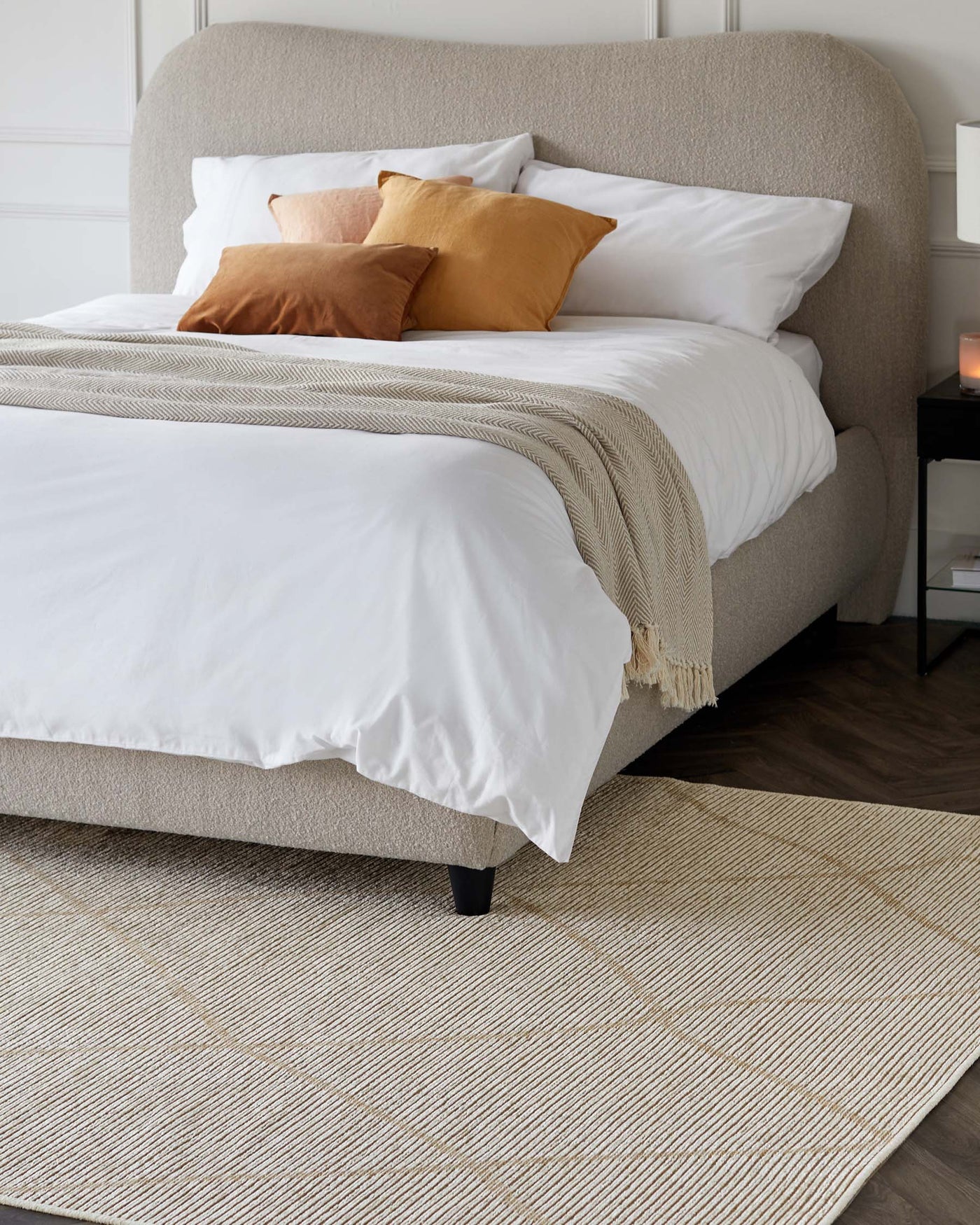 A contemporary upholstered bed with a curved headboard in a neutral beige fabric, featuring clean lines and minimalistic design. Nestled on a textured cream area rug, the bed is adorned with crisp white linens, a beige knit throw, and accent pillows in shades of mustard and terra cotta. A small black nightstand with a white lampshade partially visible, complementing the modern and cosy bedroom aesthetic.