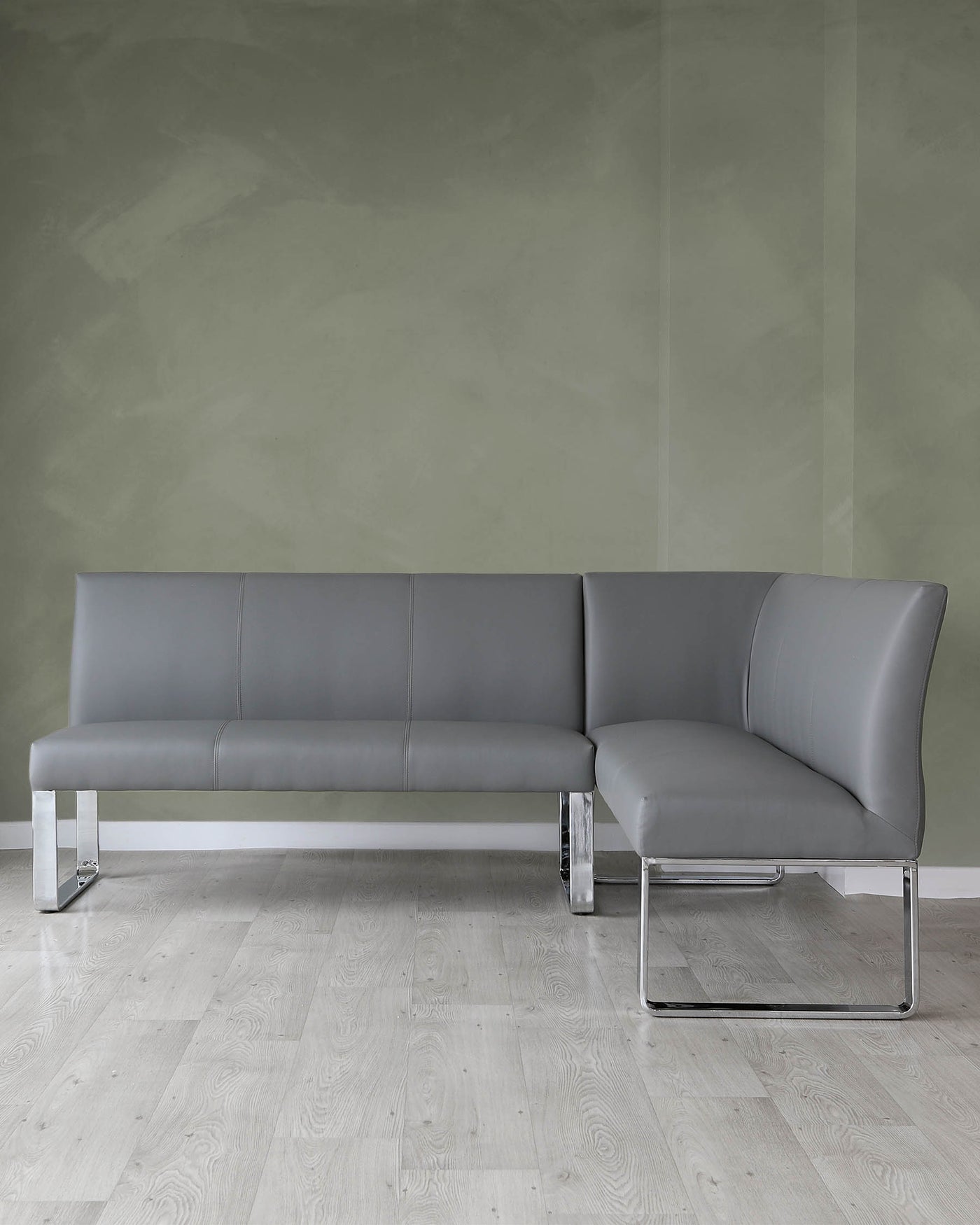 Modern grey L-shaped sectional sofa with sleek chrome legs, positioned against a textured green wall on a light wooden floor.
