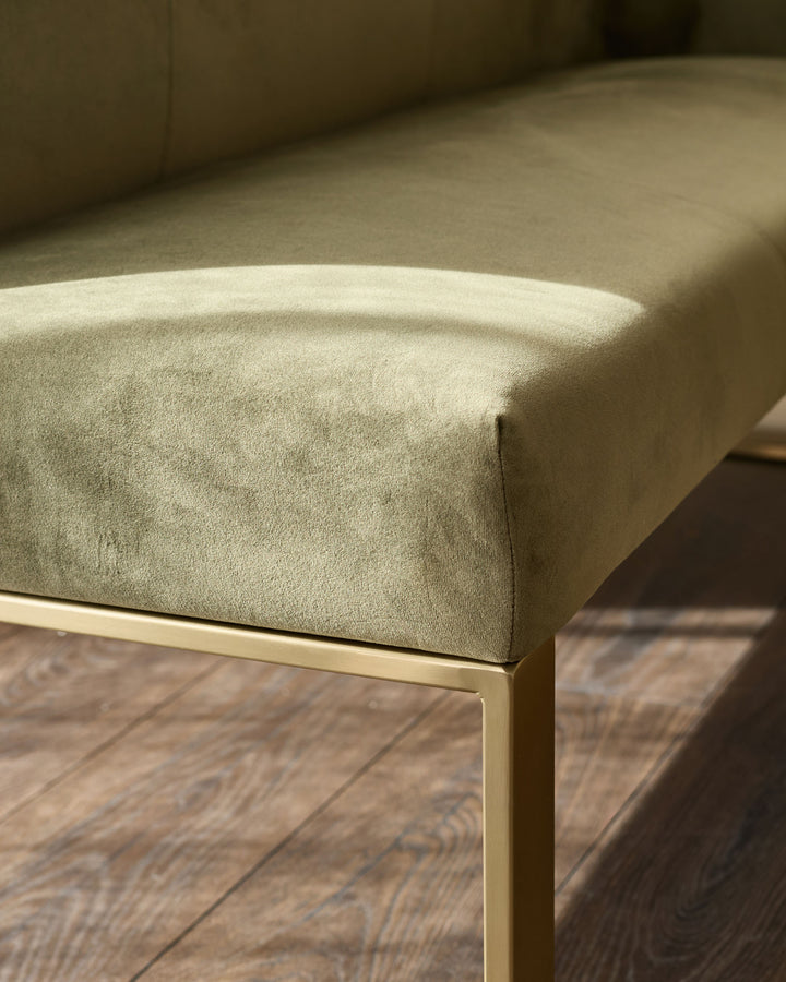 Olive green upholstered bench with brushed brass frame detail, shown from side angle on wood flooring.