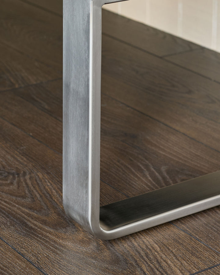 Stainless steel leg detail of Loop bench, showing brushed metal finish and curved base on dark wood floor.