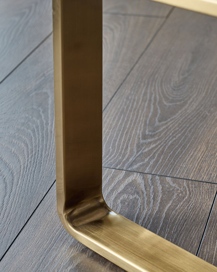 Brushed brass leg detail of grey fabric bench, angled close-up view on wooden floor.