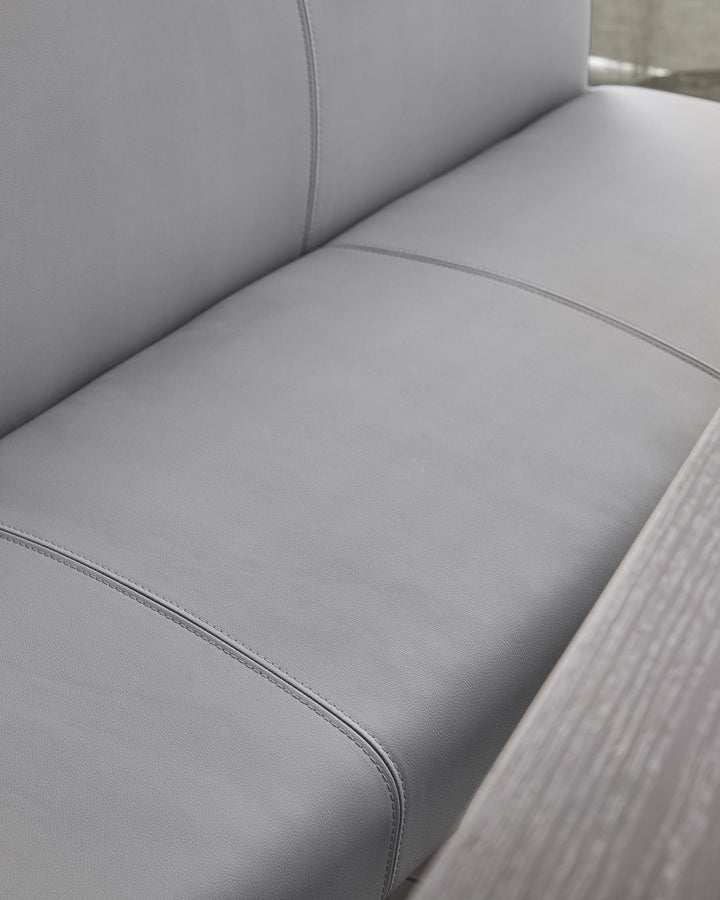 Close-up of a grey leather sofa, showcasing the texture and stitching detail, with a light-coloured wooden armrest visible.