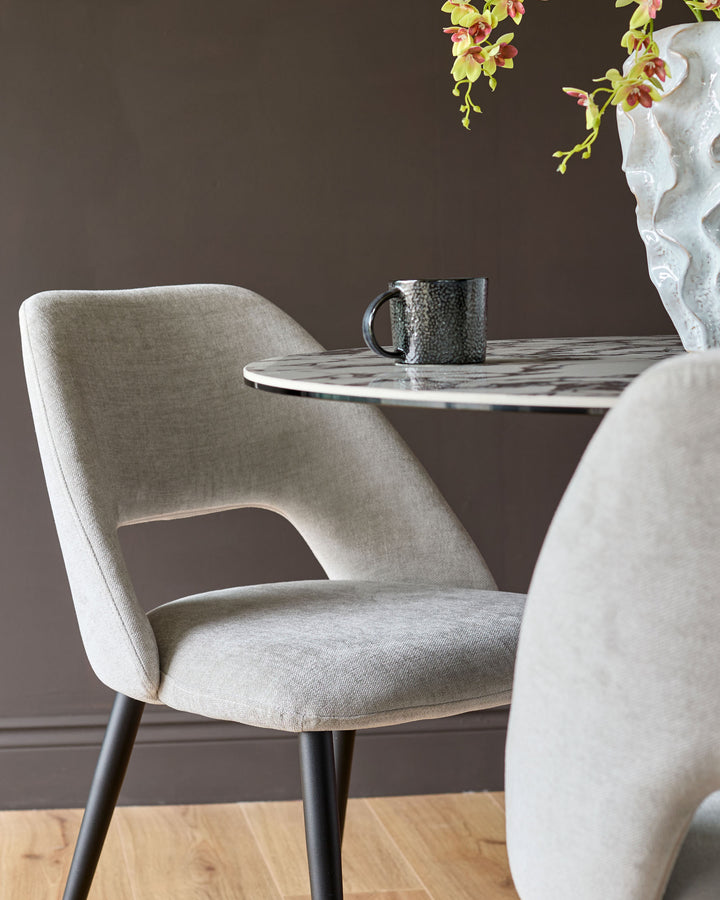 Lottie fabric dining chair in mid-grey with curved backrest and black legs, shown at a marble table with orchids.