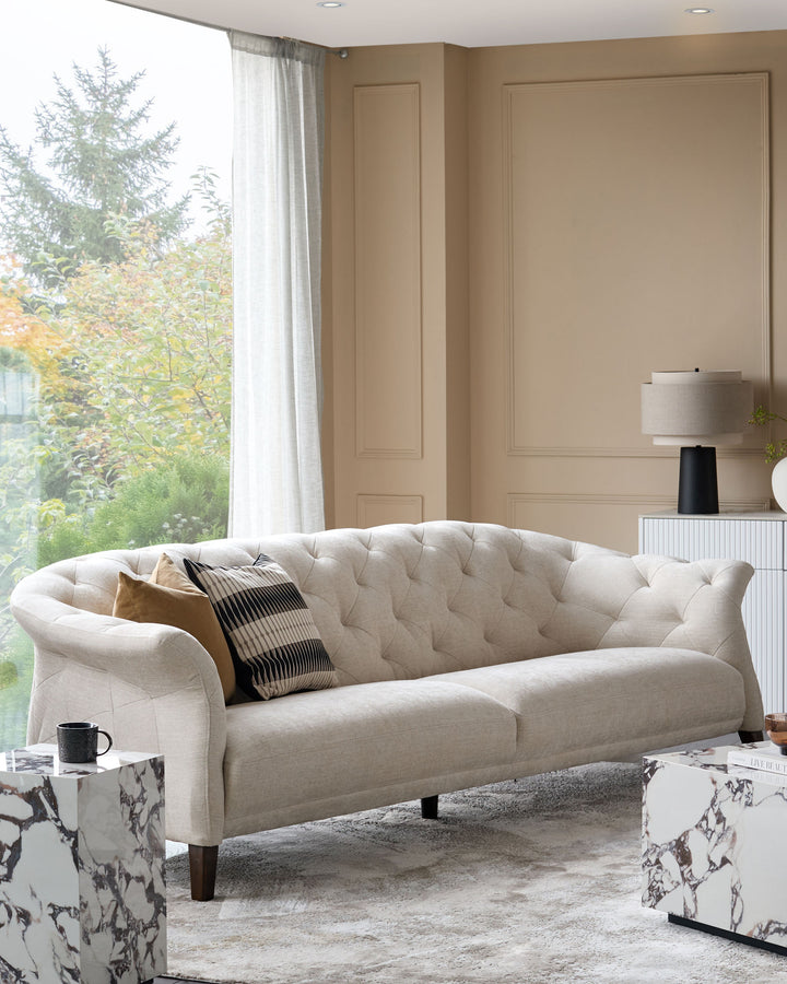 A stylish living room with a cream sofa, marble side tables, and a lamp on a white cabinet against a beige wall.